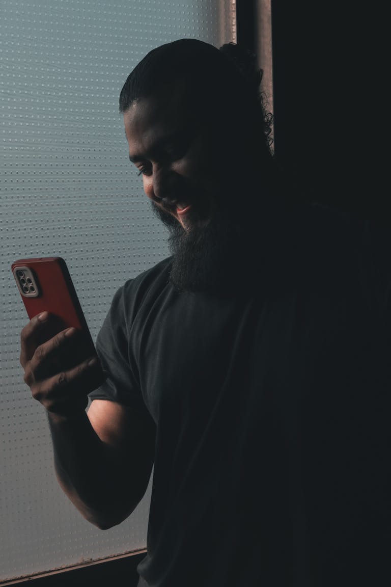 A man with a beard and ponytail smiling at his smartphone in a dark setting.