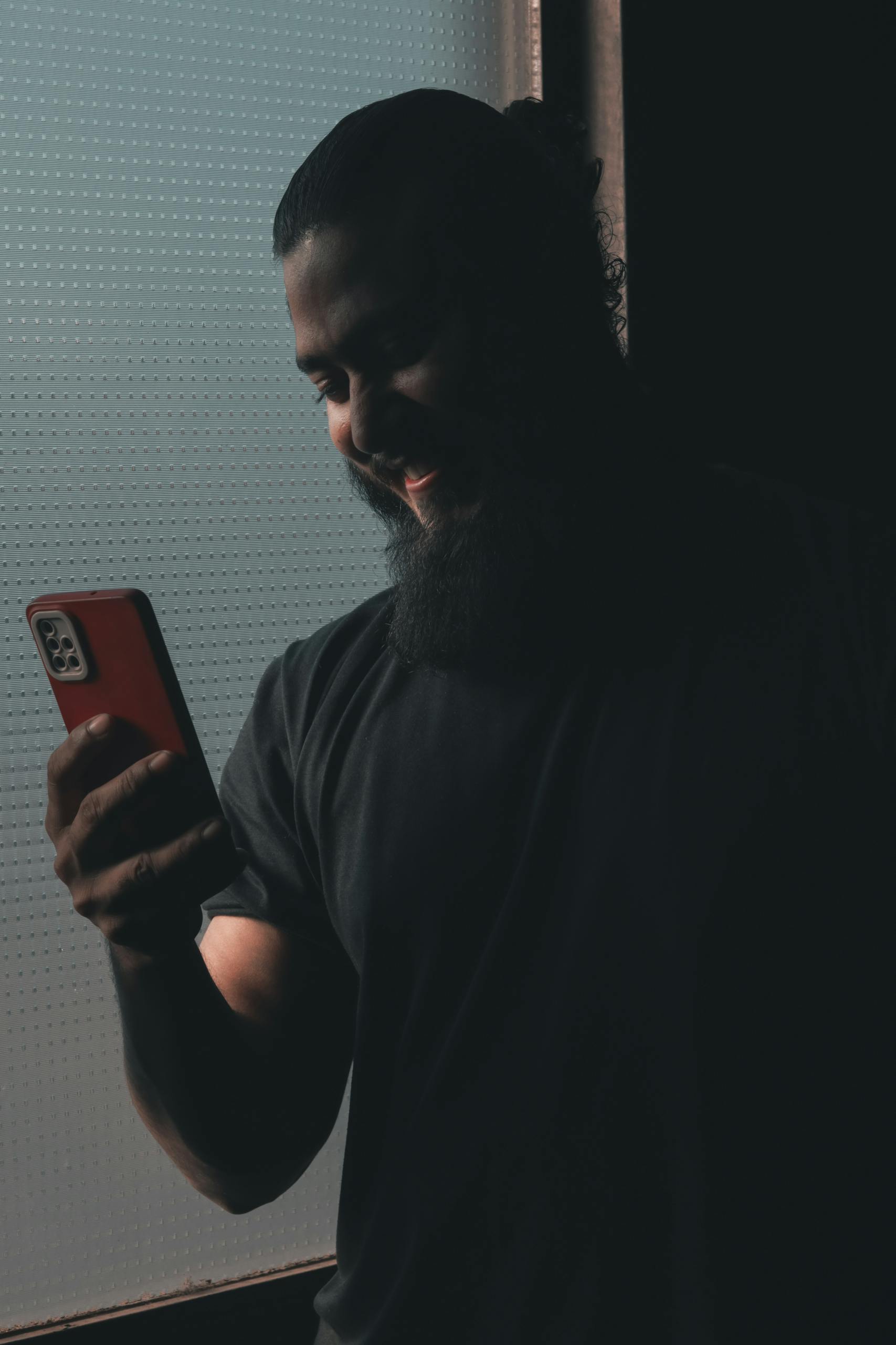 A man with a beard and ponytail smiling at his smartphone in a dark setting.