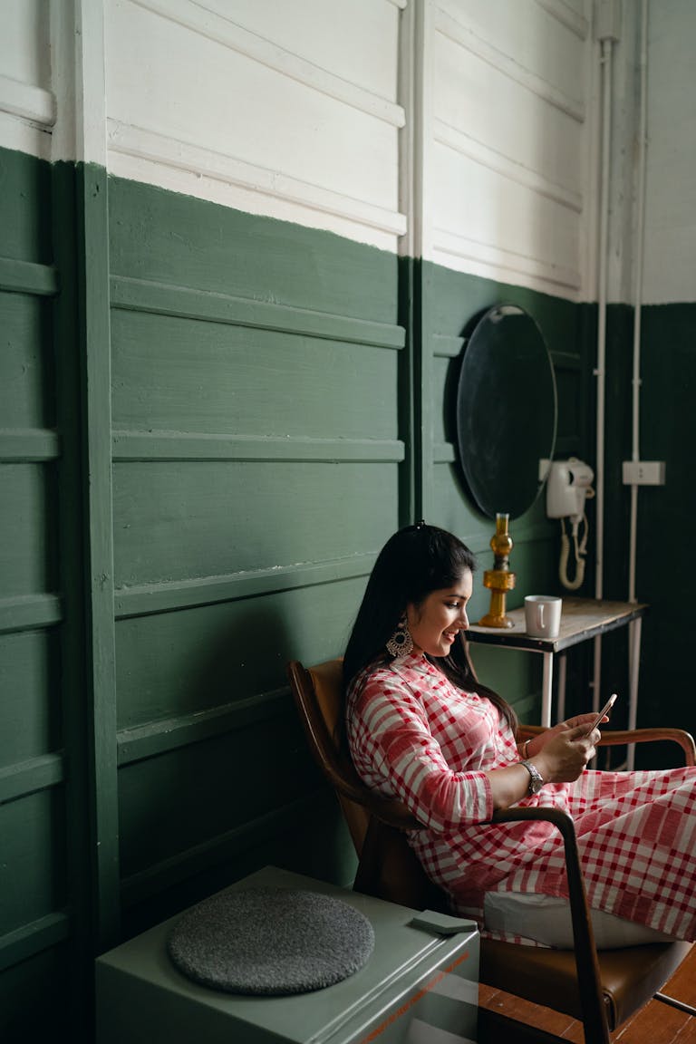 A young woman in a checkered dress relaxes and uses her smartphone in a cozy room.