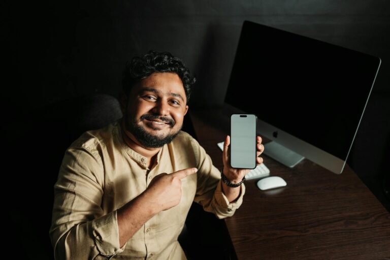 Happy man in beige shirt showing smartphone with blank screen, sitting at desk with computer.